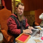 bearded man writing on laptop next to craft table covered in wooden dice trays